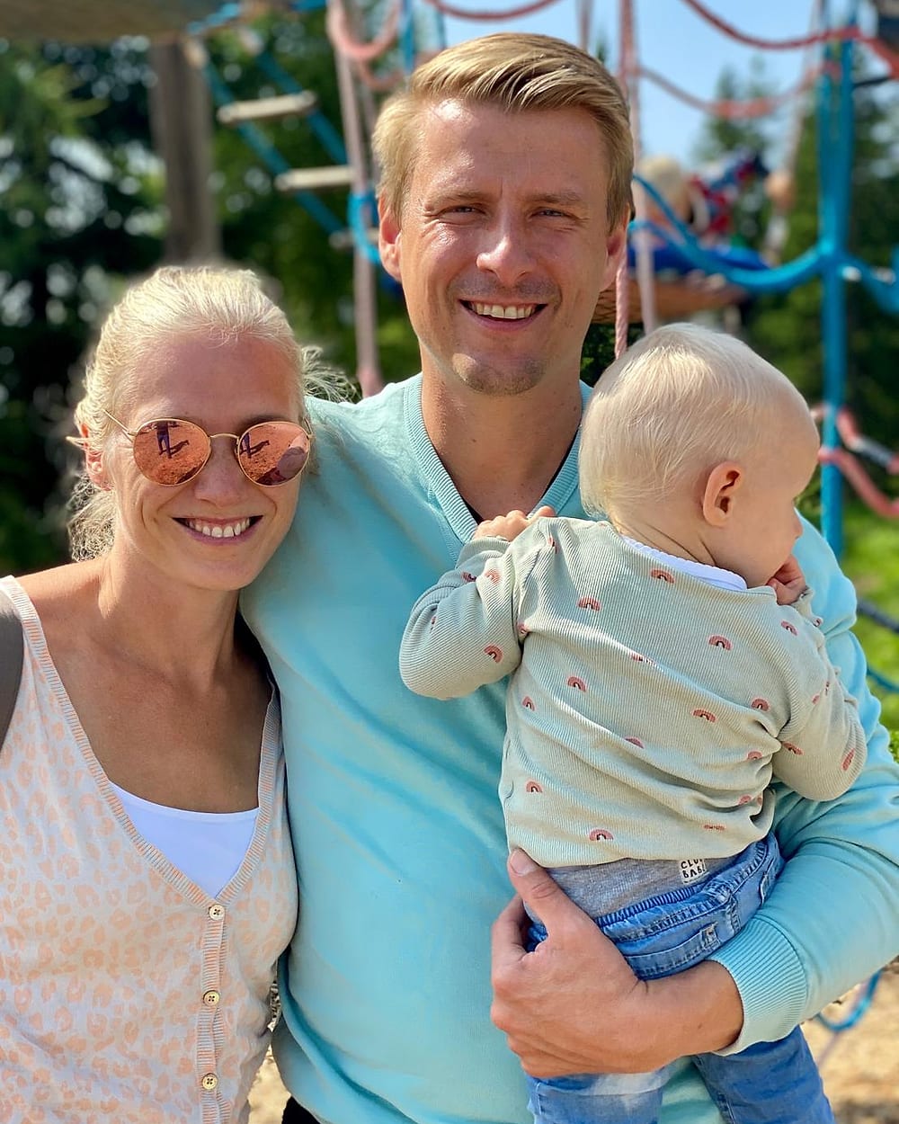 Michael Scholz lächelt in die Kamera, im Arm ein Kleinkind; neben ihm eine Frau mit Sonnenbrille, dahinter ein Klettergerüst